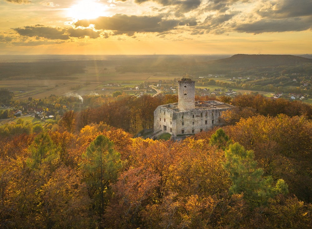 widok z lotu ptaka na zamek z 1 wieza, n aniebie przez chmurę przebija się słońce wokół drzewa zielono - złote kolory, zamek jest na wzgórzu